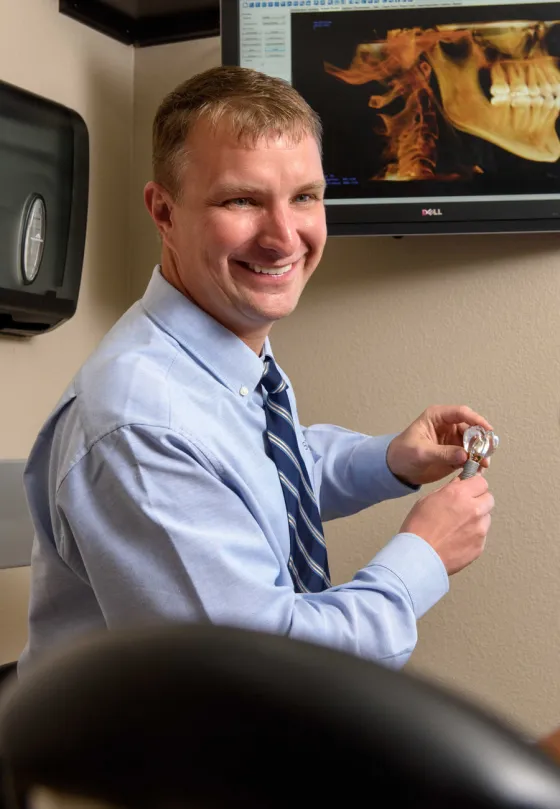 Dr. Beck, in shirt and tie, holding a dental implant model with a 3D dental scan on the screen behind him.