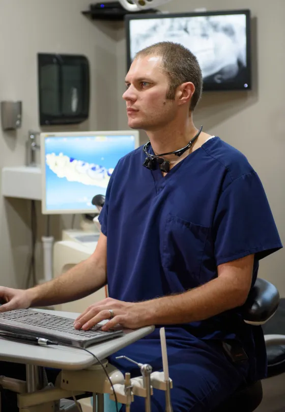 Dr. Zitterkopf in scrubs with dental loupes, working at a treatment-room computer.