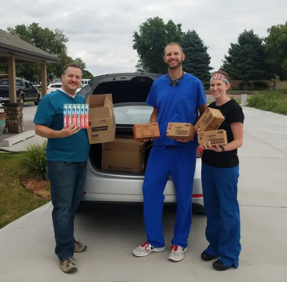 Three staff members in scrubs loading boxes of donated toothbrushes into a car.