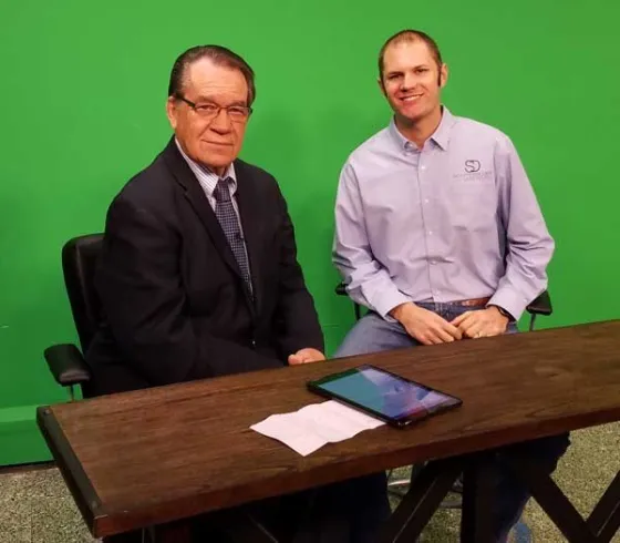 Two men seated at a news desk in front of a green screen.