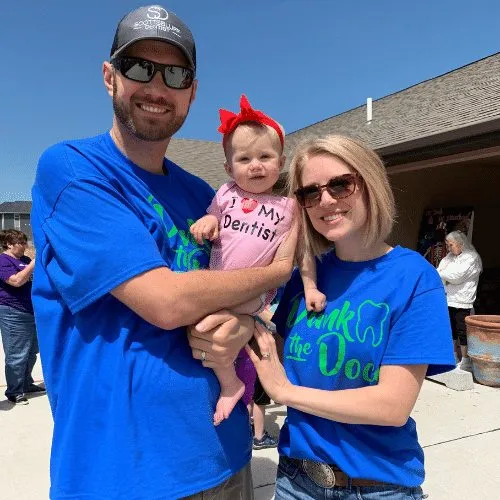 A man in a Scottsbluff Dentist shirt holding a baby beside a colleague at an outdoor event.