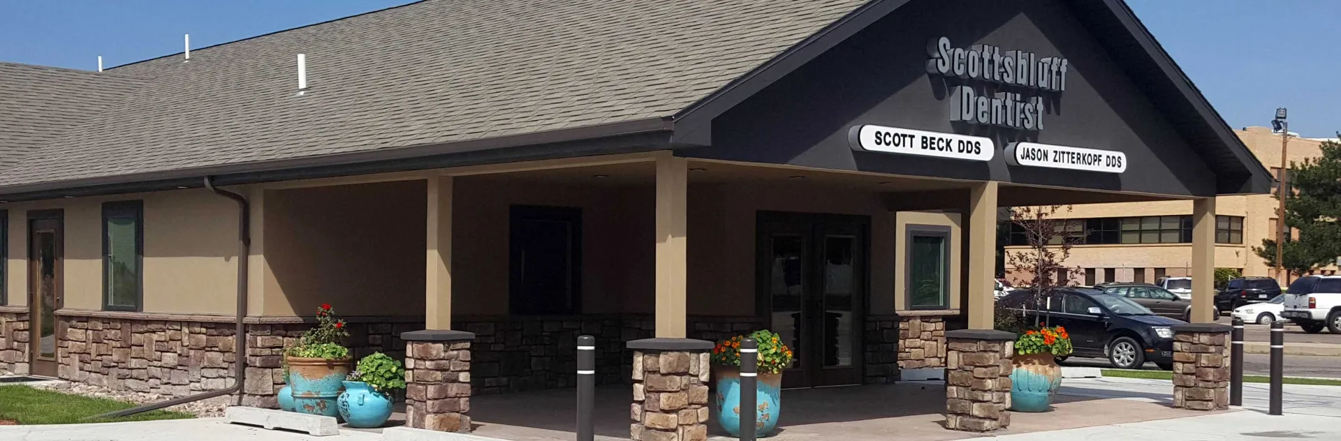 The covered entrance of the Scottsbluff Dentist office on a summer day, with signage for Scott Beck DDS and Jason Zitterkopf DDS and flowering planters along the walkway.